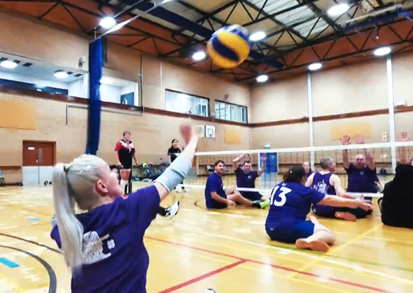 A female sitting volleyball player serves the ball towards the opposing side of the net, whilst her team mates and opposition look on ready to play, the setting is a standard sports hall court indoors.