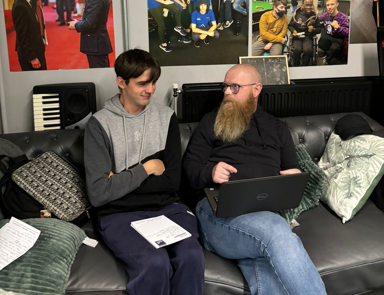 A younger member of the Academy sits on a sofa in conversation with a man who has a laptop on his lap