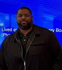 Lorn smiles at the camera, wearing a black polo shirt and jacket The background shows a blue digital display.
