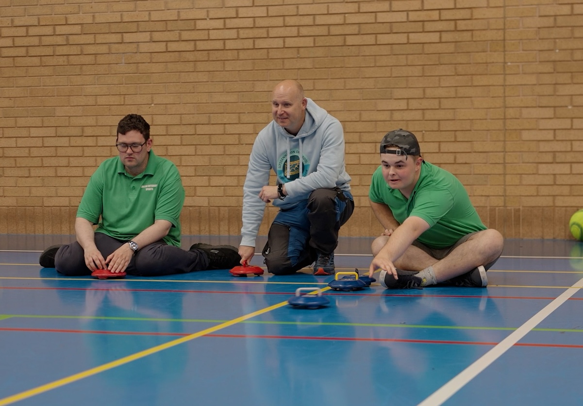 3 partcipants sat on the floor of an indoor sports hall, playing floor curling.