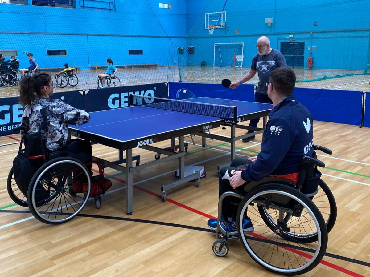 Wheelchair users play table tennis in an indoor sports hall, supported by a coach in an accessible and inclusive setting.