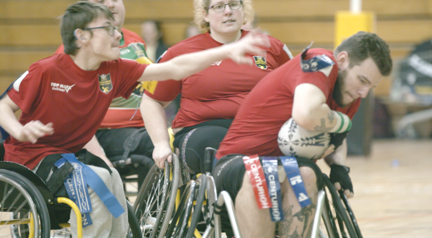 2 wheelchair rugby league players, 1 attempting a tackle and the other ducking with the ball