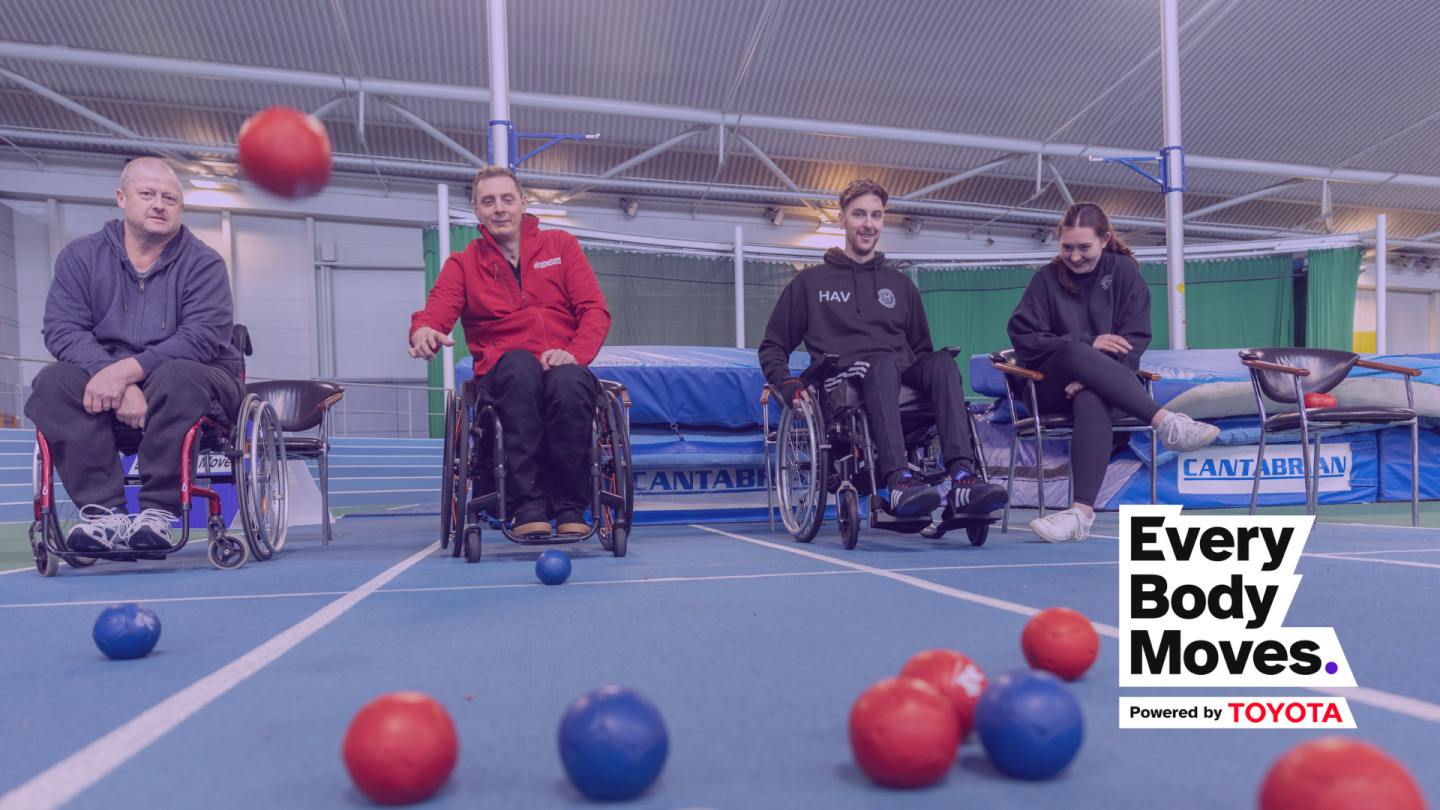 A group of 3 white males playing Boccia sat in manual wheelchairs with a white female sat next to them smiling as one of the men throws a red Boccia ball towards the camera, which is slightly blurred and in mid air as it's about to land in the middle of a set of red and blue Boccia balls.