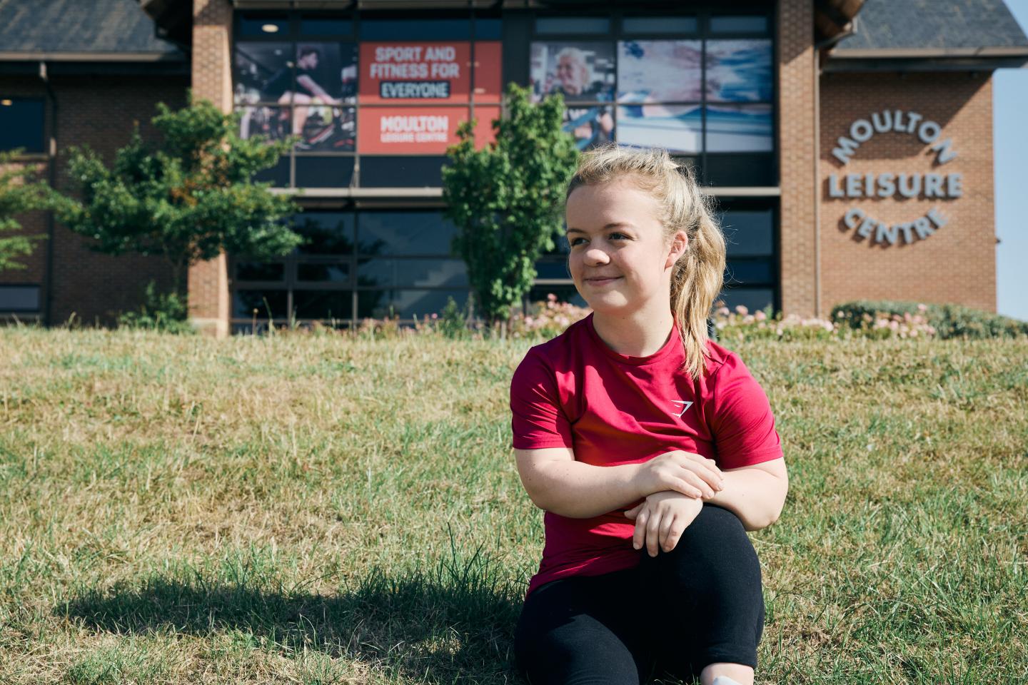 Paralympic swimmer Maisie Summers-Newton, sitting on a grassy bank outside a leisure centre.
