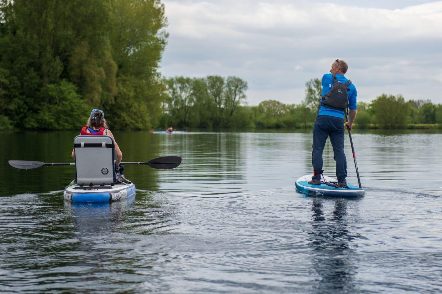 The back of a man and a woman paddleboarding on a lake. The woman is in an adapted seat whilst the man is accompanying her stood up.