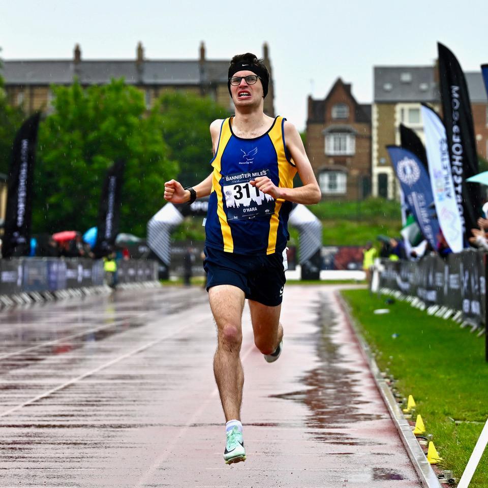 Kieran O'Hara running through a wet backdrop on a running track wearing blue running vest.