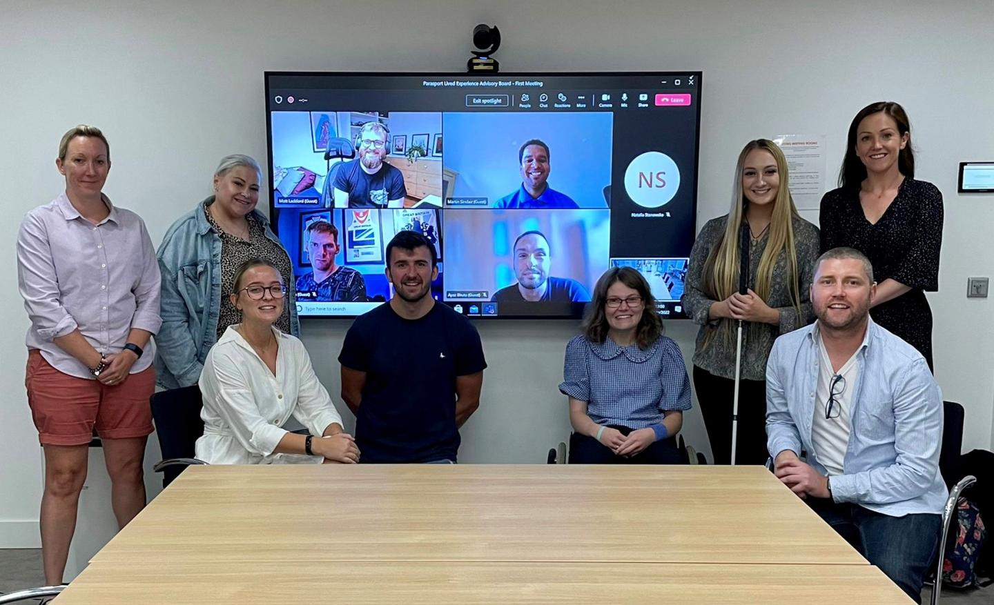 The Lived experience advisory board are pictured in a hybrid meeting with attendees in person and online pictured in a group around a large screen.