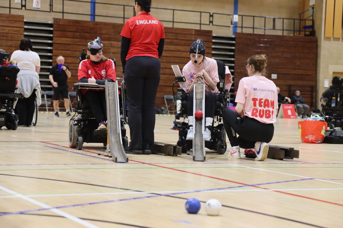 Group of boccia players mid game, sat in chairs facing the camera and using ramp launch aids