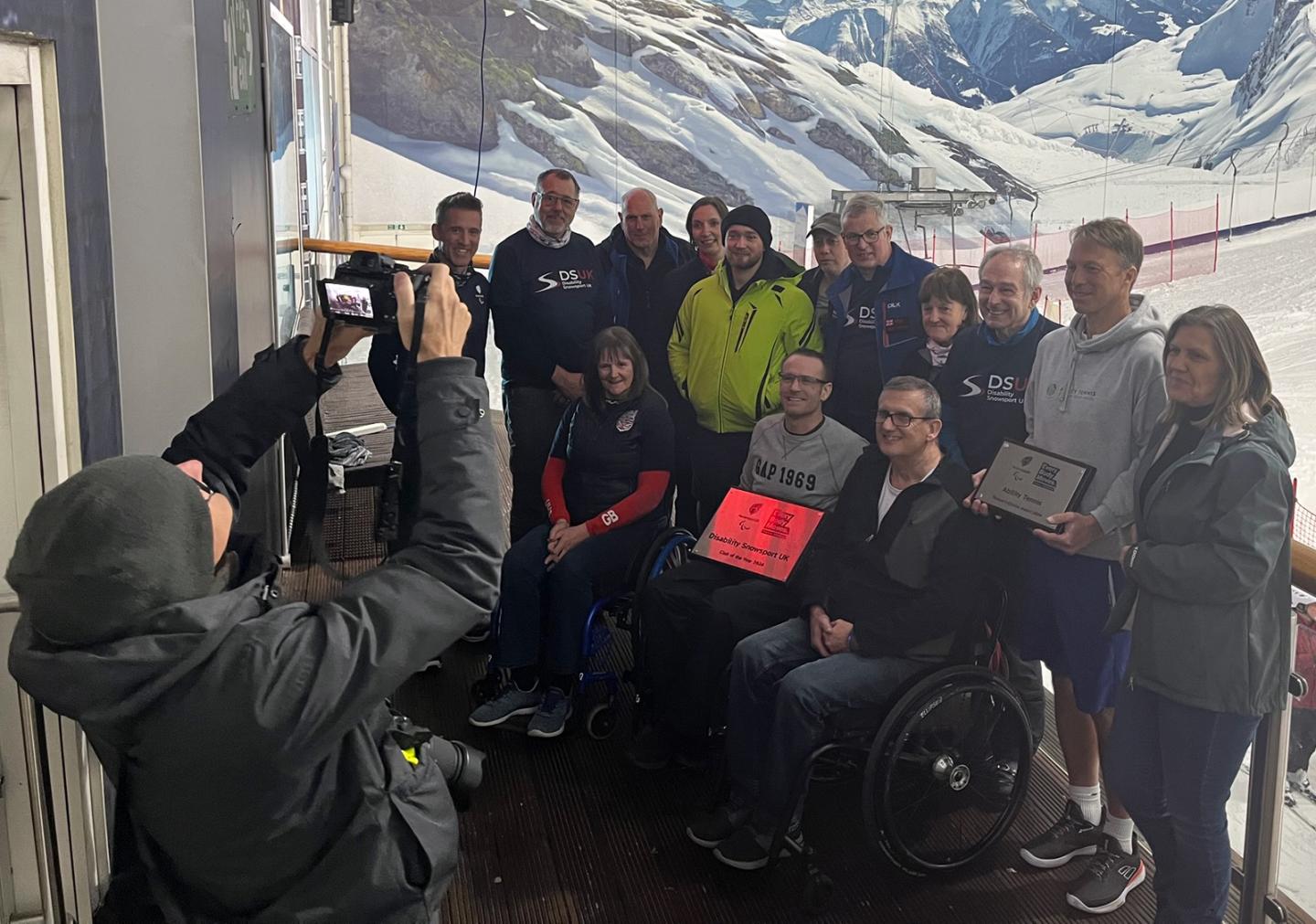 Club of the year 2024 winners DSUK, with club members on a balcony overlooking an indoor ski slope. Trophy in the middle of shot, with all looking towards a photographer taking a photo of them.