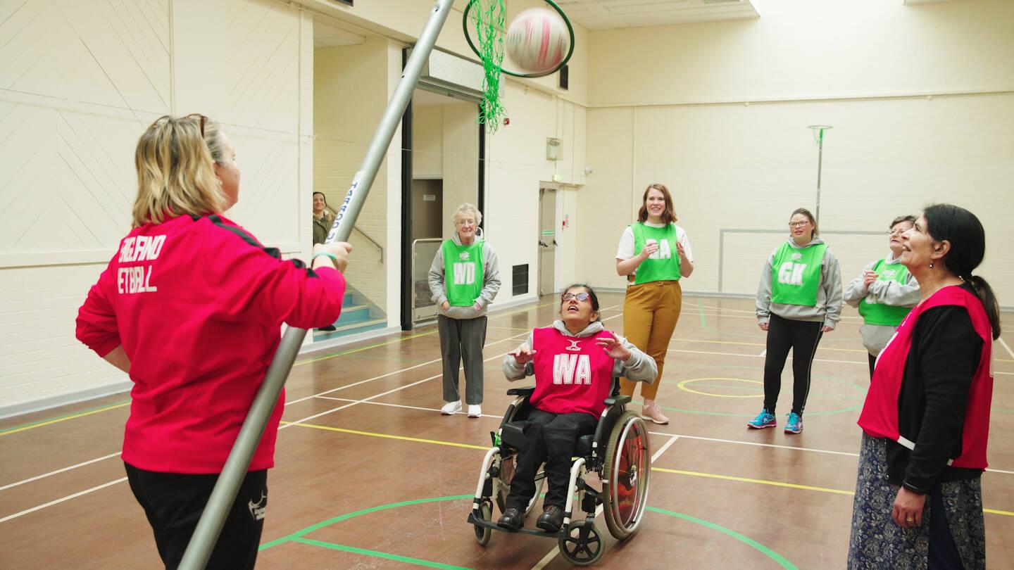 Inclusive netball session on an indoor court. Wheelchair participant taking a shot at a net being held by a coach in the foreground