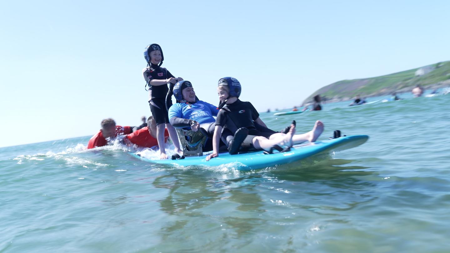 A group of children and a man using adaptive equipment enjoying an inclusive surfing session on a sunny day, supported by volunteers in the sea, with a scenic coastline in the background.