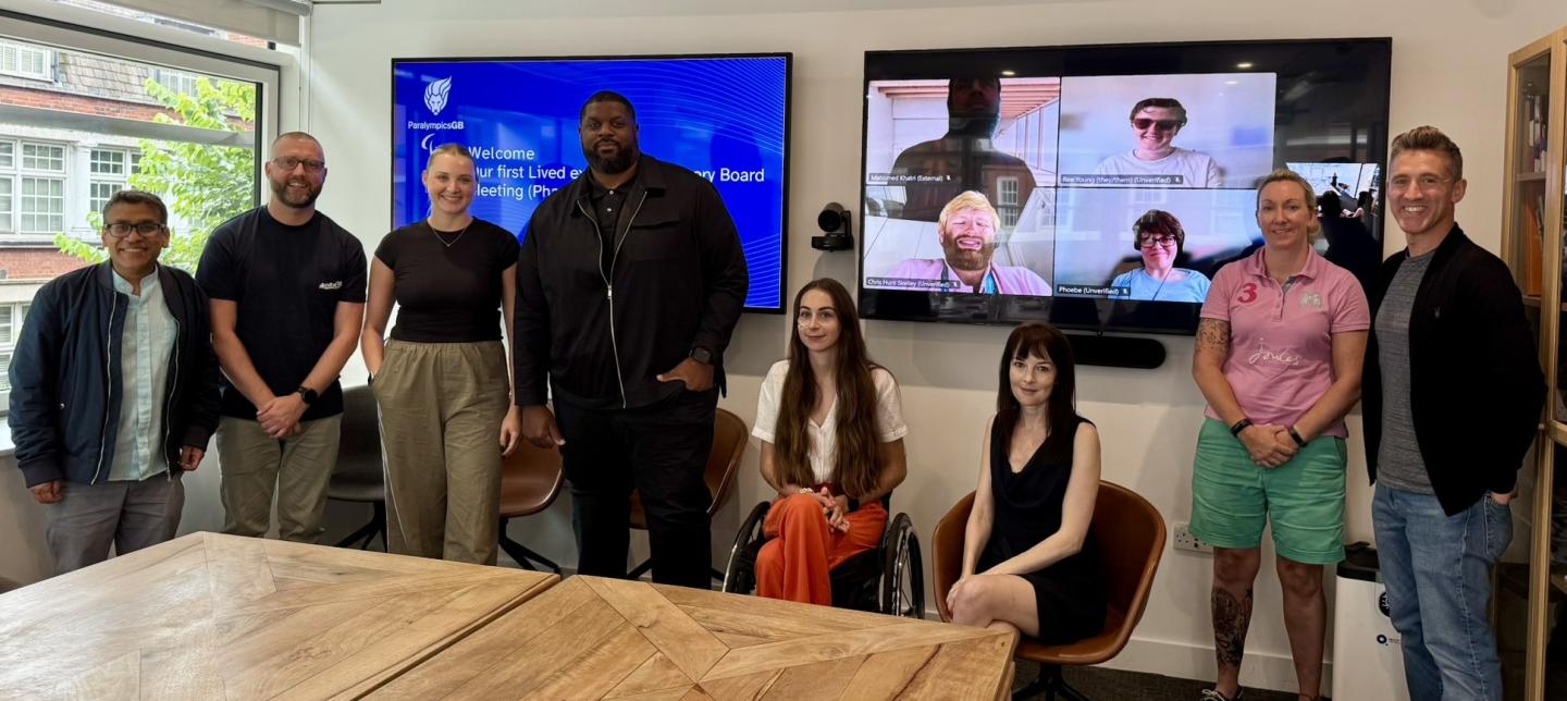 A group photo from the first in-person meeting of ParalympicsGB’s Social Impact - Lived Experience Advisory Board (Cohort 2). Six advisory board members stand or sit in a meeting room alongside two ParalympicsGB staff members, with four others joining via video call displayed on a screen behind them. The two large screens shows a ParalympicsGB welcome slide and video call participants. The group is diverse in age, gender, ethnicity, and disability. The atmosphere is friendly and collaborative.