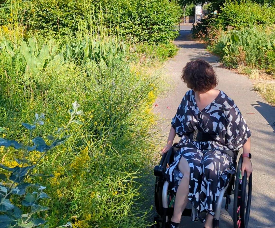 Meg, sitting in her wheelchair surrounded by lush green plants and foliage, in an inner city park 