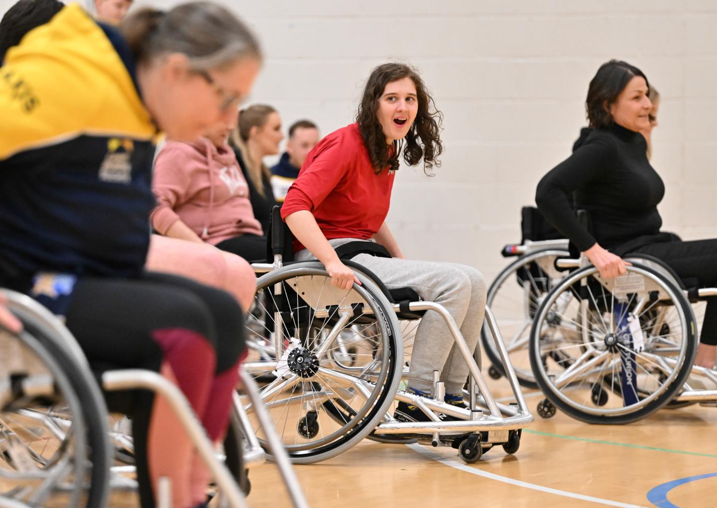 A young woman in a red top and grey tracksuit bottoms takes part in a wheelchair rugby league session on an indoor court. She looks animated and engaged as she pushes her chair forward, surrounded by other participants also using sports wheelchairs. A group of people watch from the sidelines in the background.
