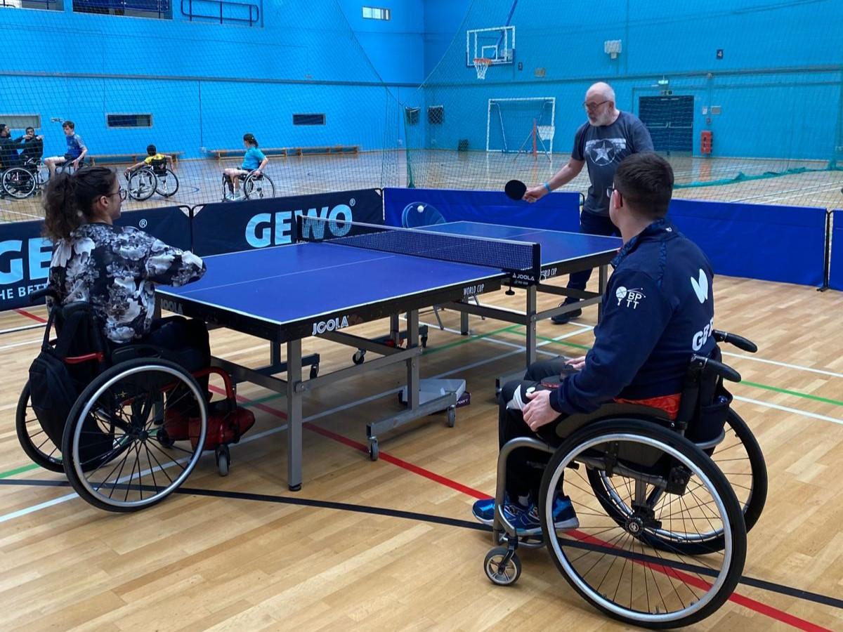 Wheelchair users play table tennis in an indoor sports hall, supported by a coach in an accessible and inclusive setting.