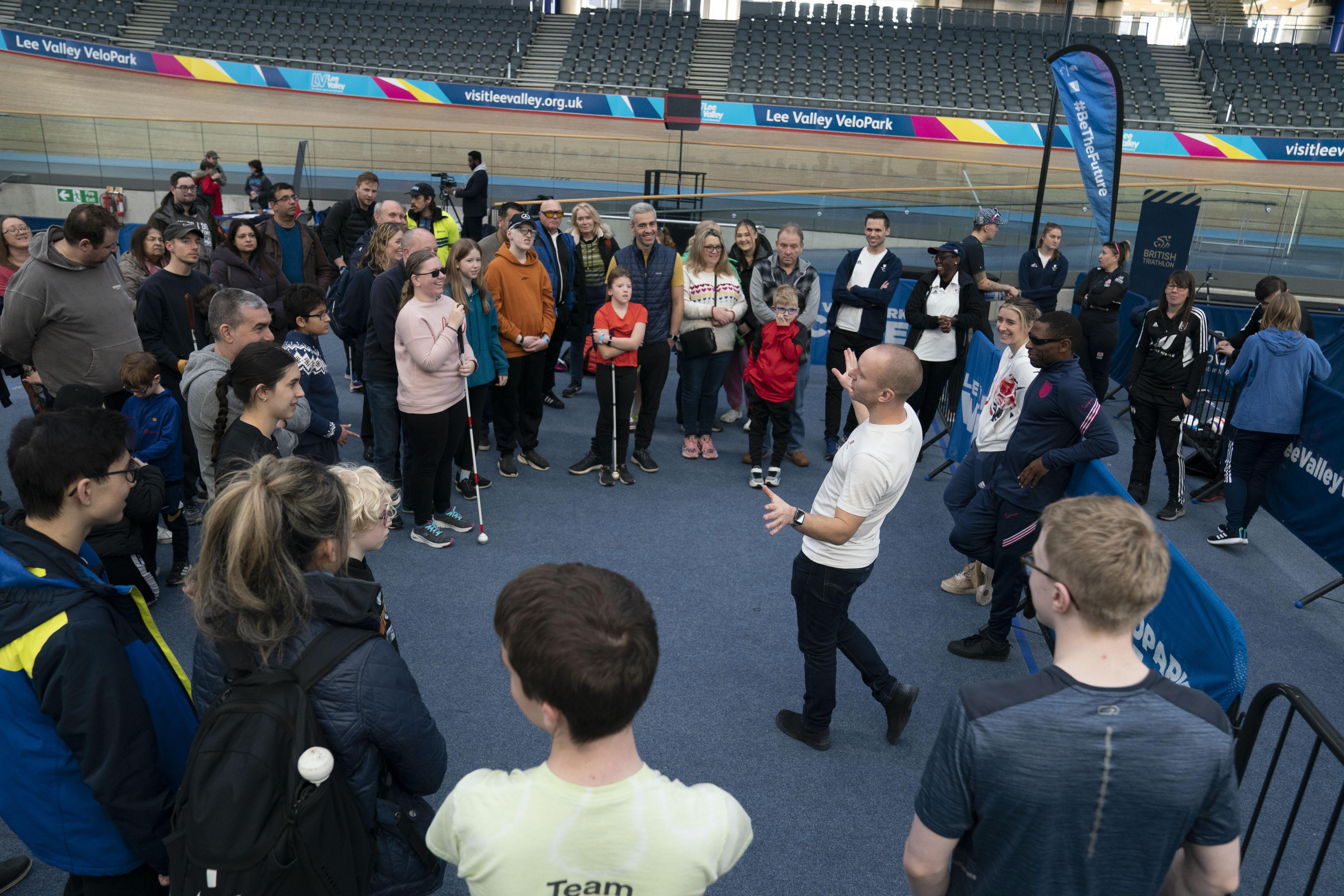 A group gathers round our Paralympic potentials lead and former Paralympic athlete Ben Quilter who is delivering a briefing ahead of a previous give it a go day. The backdrop is an indoor velodrome.