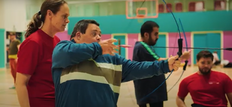 A female coach with a male club member shooting an archery bow. In the background another coach looks on with another club member.