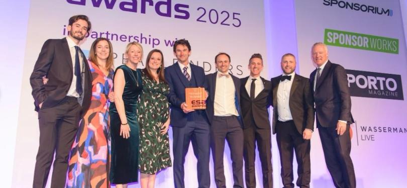 A group of nine people dressed in formal attire stands on stage at the ESA Awards 2025, smiling and celebrating. The central figure holds a wooden award plaque, and the backdrop displays event sponsors and logos, including "ESA Awards" and "Wasserman Live."