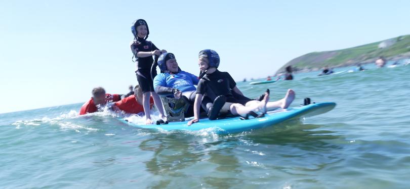 A group of children and a man using adaptive equipment enjoying an inclusive surfing session on a sunny day, supported by volunteers in the sea, with a scenic coastline in the background.