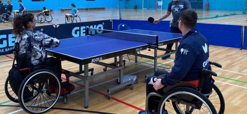 Wheelchair users play table tennis in an indoor sports hall, supported by a coach in an accessible and inclusive setting.