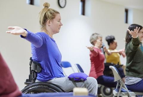Wheelchair user with blond hair tied up in a top knot wearing a blue shirt extending her arms out to the side with a sandbag on her lap to help ground her in the yoga pose , other students, one in a red top, one in a yellow top an one in a green top all mimic the pose in the background 