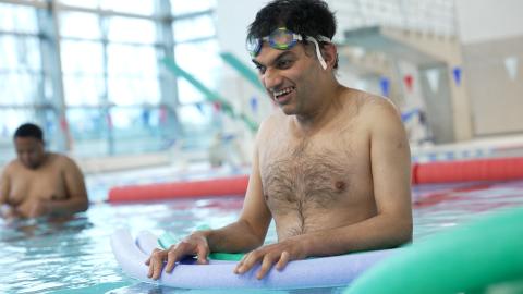 male adaptive swimmer, wearing goggles while stood in the pool and holding on to flotation aids