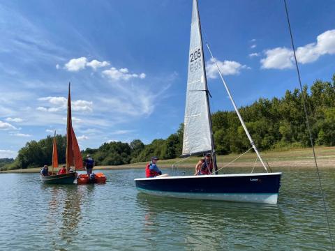 2 small sailing boats with triangular sails at full mast. Heading from left to right of picture against blue skies, with lush green trees across the nearby shore and calm waters.