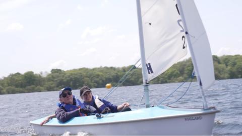 Small 2 person sailing boat containing father and daughter sailors, Ivor and Kathryn. They are sat low in the board, with triangular sail full, and the boat is heading from left to right of photo