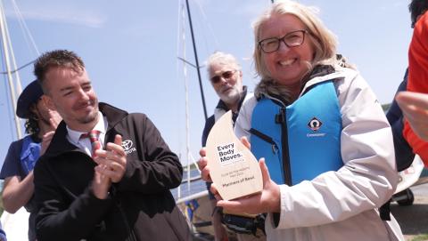 Club chair, Carolyn Howden, holding the Every Body Moves Club of the Month award. Attendees around her are applauding against a backdrop of sailing boats.