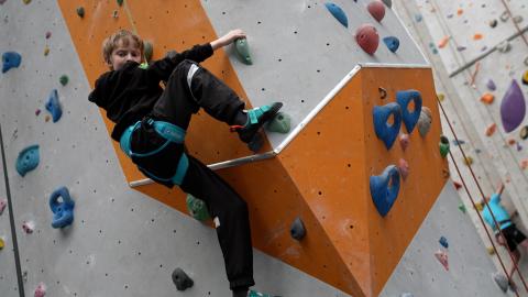 A young paraclimbing boy mid climb across and overhanging section. Both his arm are straight in front across his body, grabbing holds on top of the overhang His right knee is bent and right foot also on a hold above the overhang, left leg is straight and using a hold below the overhang. He is looking back down towards his instrcutor.