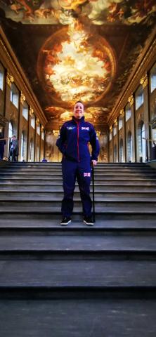 Stacey Denyer - Dressed in her blue Invictus Games athlete uniform. Using a cane while stood on a set of steps leading to a very regal looking hall, with pillared arches each side and ornate painted ceiling.