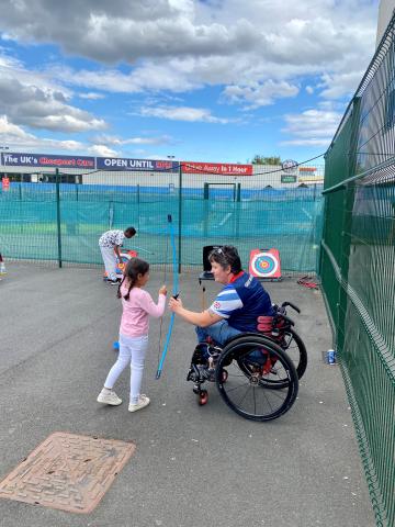 Wheelchair user teaching a young girl how to shoot a bow, with the soft archery range set up in a school playground.