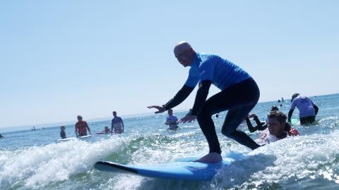 Visually impaired male surfer standing up on a surfboard for the very first time. He is stood sideways on the board with a low and balance posture, arms spread out low in front of him. The nose of the board is lifted as it rides the wave in and the water is white and foamy as it breaks either side. An instructor is in the water holding onto the back of the board, aiding with steering and giving instructions to the surfer.