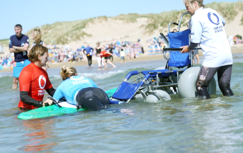 Female wheelchair user laying on a surf board after being helped from a beach accessible wheelchair into the water. A male coach in red top is kneeling in the shallows holding her surfboard steady, another volunteer is stood and supporting the wheelchair.