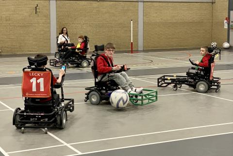 4 powerchair football players, mid pass and tackle during a game at an indoor sports hall.