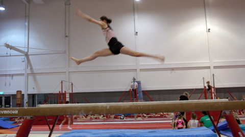 Young female adaptive gymnast performing a splits jump on the beam.