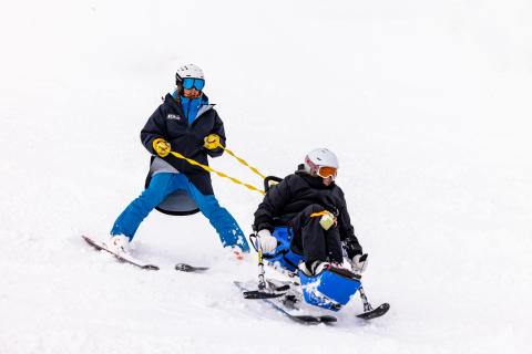 A sit skier enjoying a run on a snowy mountain, with the sit ski tethered and being supported by a guide skiing behind them.