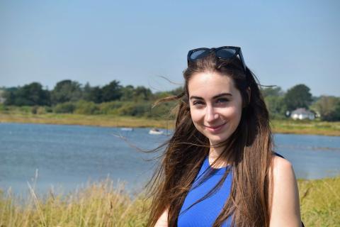 Bethany, a young woman with long straight dark hair, smiles at the camera. She’s wearing a blue sleeveless top and sunglasses resting on her head. Behind her is a sunny riverside scene with trees, grass, and moored boats in the background.