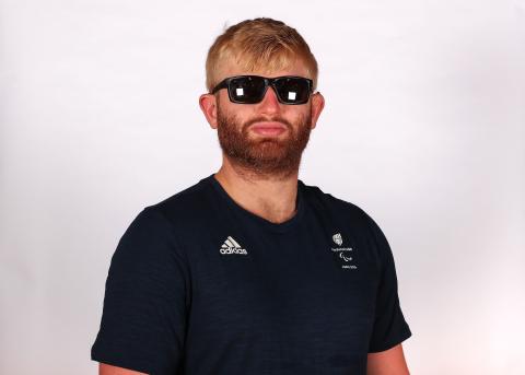 Chris, a man with short fair hair, a trimmed beard, stands facing the camera. He’s wearing dark sunglasses and a navy ParalympicsGB t-shirt with the adidas logo. The background is plain and light-coloured.