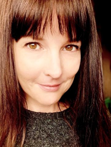 Ellie, a woman with long straight dark hair and a fringe, smiles at the camera. wearing a dark textured top.