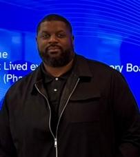 Lorn smiles at the camera, wearing a black polo shirt and jacket The background shows a blue digital display.