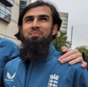 Mahomed smiles with his mouth closed, wearing an England cricket team jacket with the team crest and Castore logo. He has a full dark beard and neatly combed hair. A hand rests supportively on his shoulder, and buildings are visible in the background.