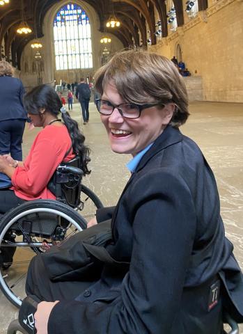 Phoebe smiles warmly while seated in Westminster Hall at the UK Parliament. The historic hall’s arched ceiling and large stained-glass window create a dramatic backdrop. 