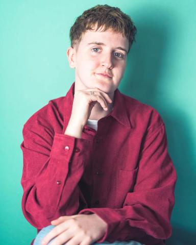 Ree sits in front of a turquoise background, resting their chin on one hand and smiling. They wear a loose-fitting red shirt over a striped top, with short, wavy hair neatly styled.