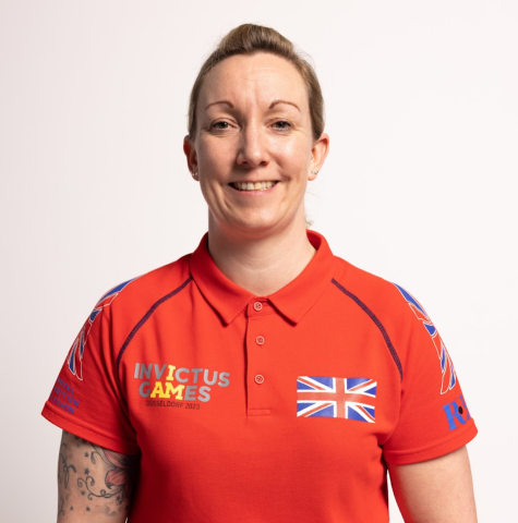 Stacey smiles at the camera wearing a red Invictus Games polo shirt with Union Jack flags on the chest. She has light brown hair tied back and tattoos on her right arm, pictured standing against a plain light background.