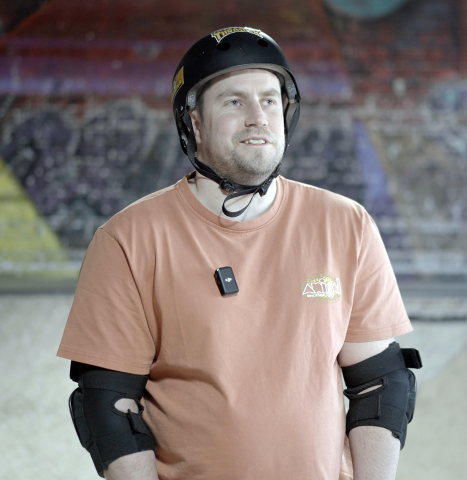 VI skateboarder, Tom, wearing a helmet and t-shirt at an indoor skatepark. Looking slightly off camera and smiling.