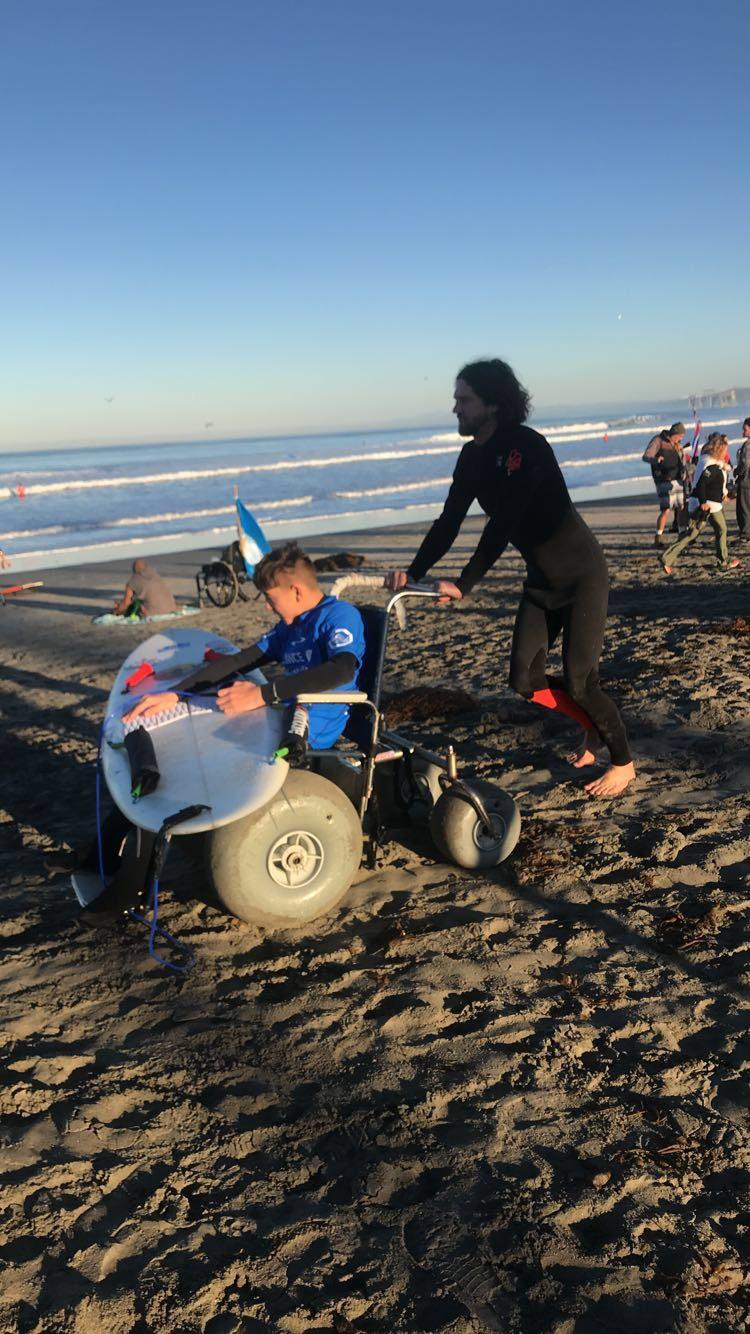 Coach Ben with prone surfer Ethan showing his beach wheelchair and prone surfboard 