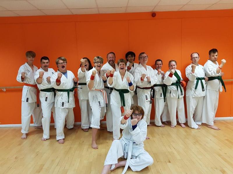Group of disabled and non-disabled people laughing and wearing karate suits and their new green belts. One person is stting on the floor