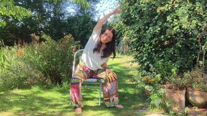 AYL teacher Paola is sat in a chair with her right arm stretching over her head, as she bends her torso, in a nice side stretch. Shes in a beautiful green garden and is wearing a white tshirt and colourful baggy trousers 