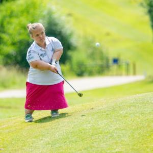 A female person of short stature playing golf, having just hit the ball